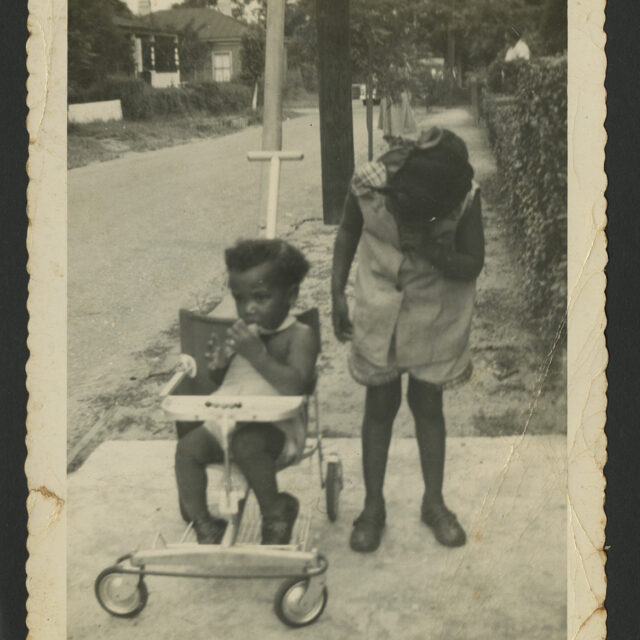 A black-and-white photograph of young Venus Jones standing next to her younger brother, Albert on their driveway. Venus is covering her face with her hand, and Albert is sitting in a stroller.