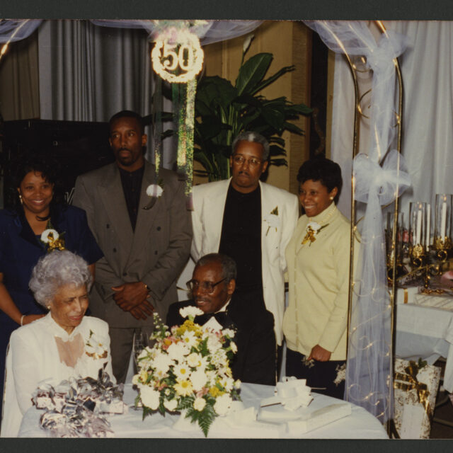 Venus Jones' parents sit at a table together and smile. Their children, Vicki, Culbert, Albert, and Venus Jones stand behind them at their celebration for their 50th wedding anniversary.