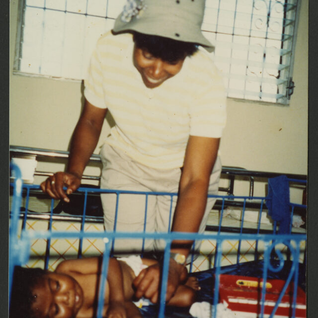 Venus Jones smiles down at her adopted son, Odell, as she meets him in a nun-operated hospital in Haiti. Odell is lying in a crib.